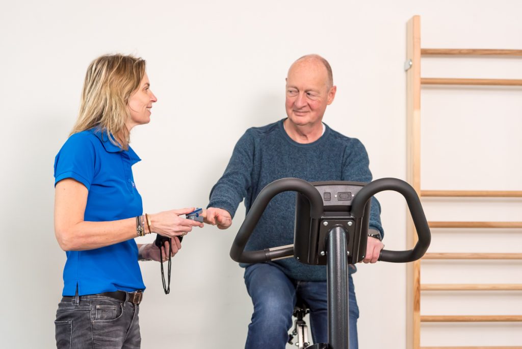 Man in zijn pensioen traint op een hometrainer onder begeleiding van een fysiotherapeut.
